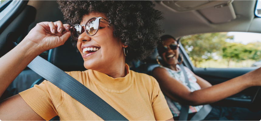 Two smiling women driving in a car while wearing their seatbelts. The woman in the passenger seat is wearing sunglasses and looking out the window.