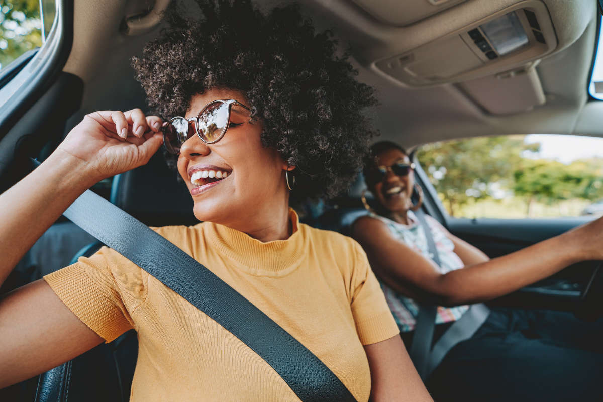 Two women enjoying a car ride on a sunny day. The woman in the passenger seat, wearing a mustard yellow top and sunglasses, is laughing while adjusting her glasses. The driver, also smiling, is wearing a sleeveless top and sunglasses. Both are wearing seatbelts, and the car's interior is well-lit with natural light coming through the windows, showing trees in the background.